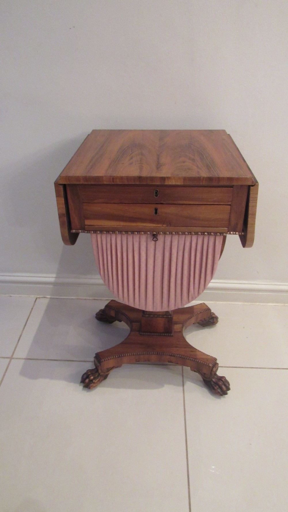 A William IV flame mahogany work table featuring drop-leaf side flaps, drawers with keyholes, a pleated pink fabric work well, and a quadruped base with carved paw feet.