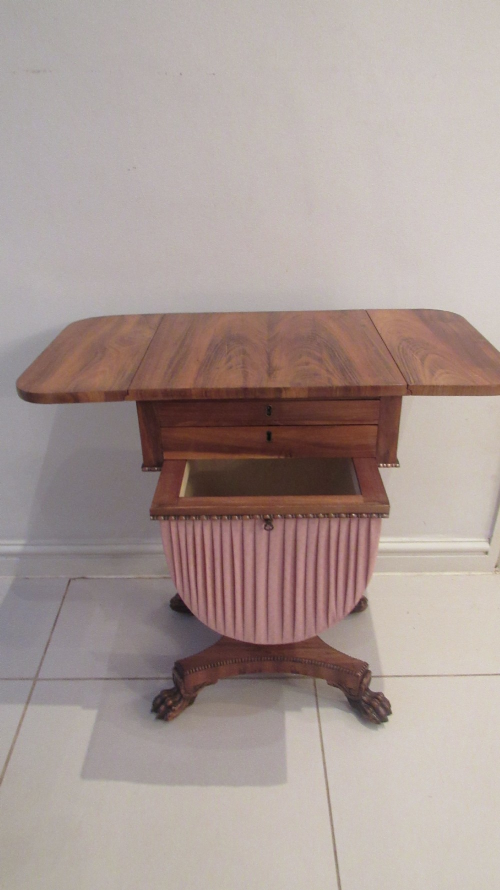 A William IV flame mahogany work table featuring drop-leaf side flaps, drawers with keyholes, a pleated pink fabric work well, and a quadruped base with carved paw feet. View III