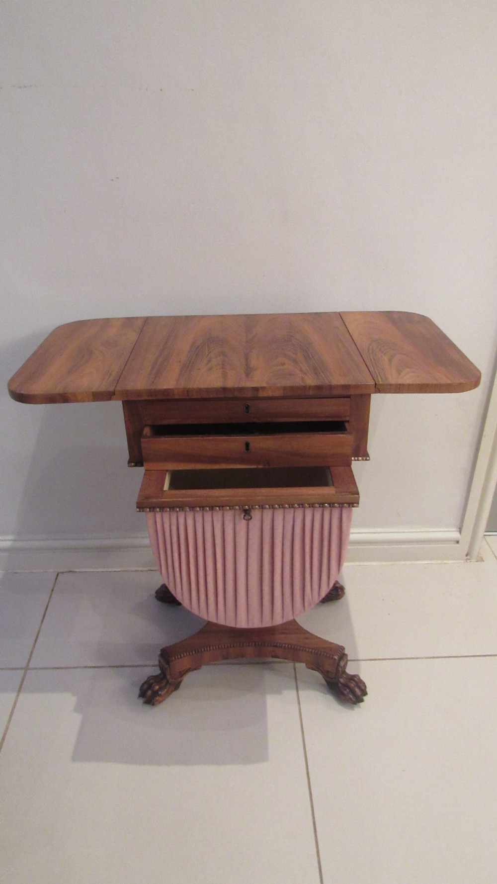 A William IV flame mahogany work table featuring drop-leaf side flaps, drawers with keyholes, a pleated pink fabric work well, and a quadruped base with carved paw feet. View V