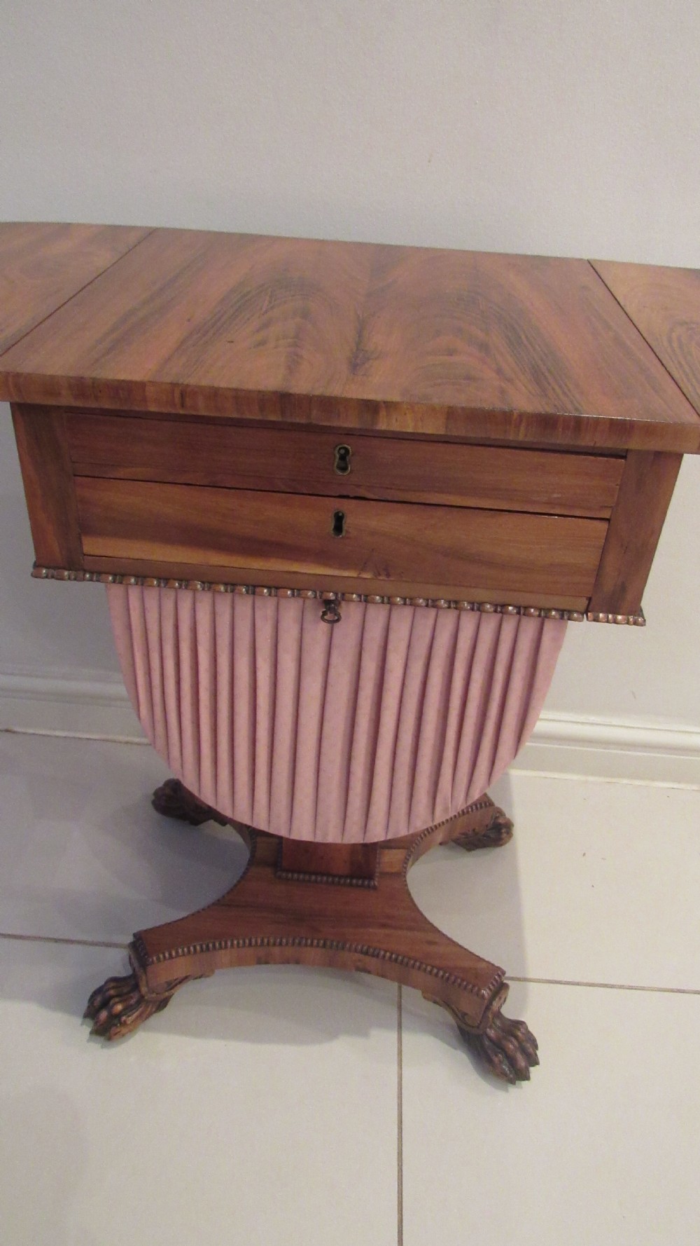 A William IV flame mahogany work table featuring drop-leaf side flaps, drawers with keyholes, a pleated pink fabric work well, and a quadruped base with carved paw feet. View XIII