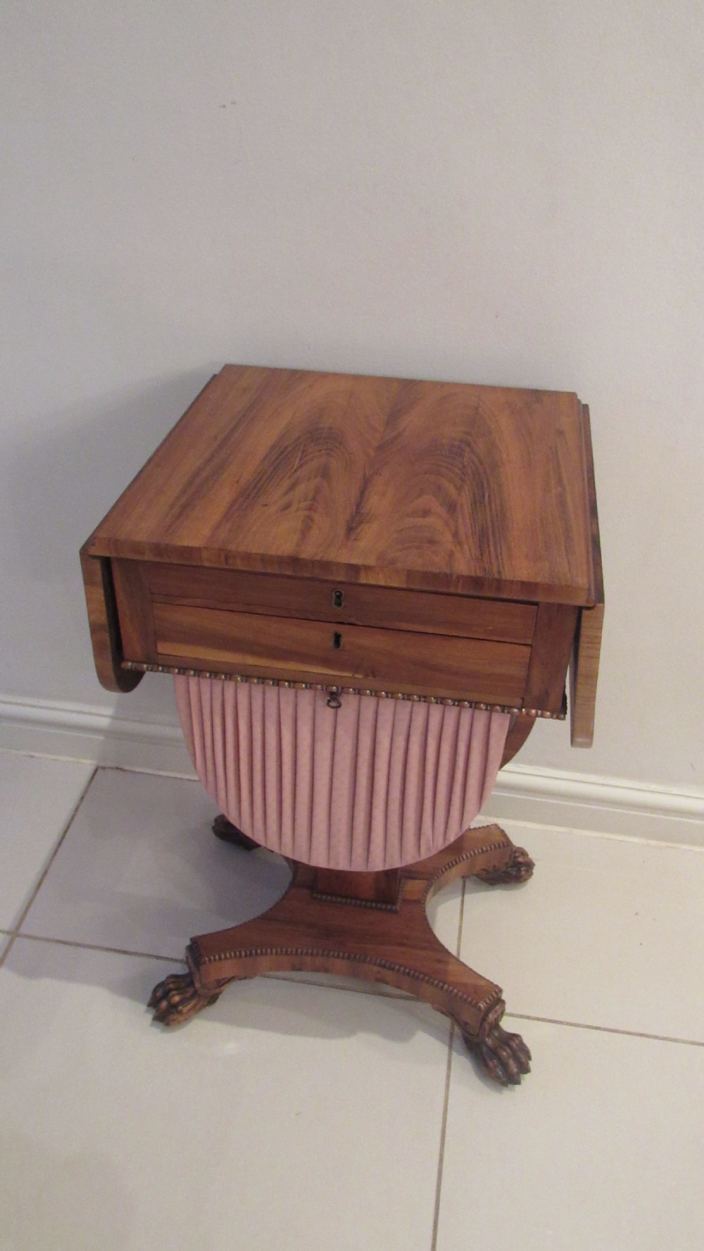 A William IV flame mahogany work table featuring drop-leaf side flaps, drawers with keyholes, a pleated pink fabric work well, and a quadruped base with carved paw feet. View XVII