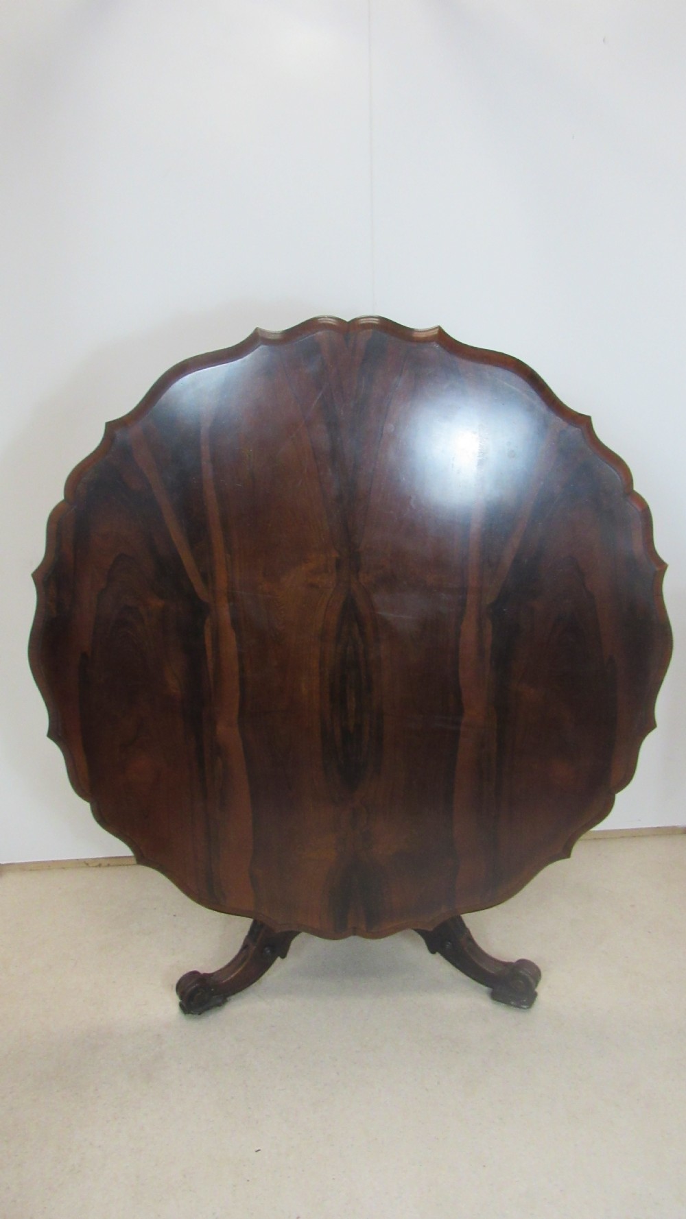 A 19th-century circular rosewood tilt-top table displayed in an upright position, featuring a shaped scalloped rim, prominent book-matched timber grain, and a carved pedestal base.