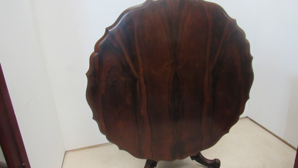 A 19th-century circular rosewood tilt-top table displayed in an upright position, featuring a shaped scalloped rim, prominent book-matched timber grain, and a carved pedestal base. View II