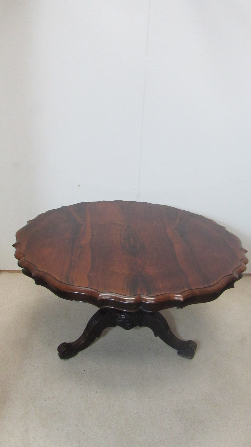 A 19th-century circular rosewood tilt-top table displayed in an upright position, featuring a shaped scalloped rim, prominent book-matched timber grain, and a carved pedestal base. View IV
