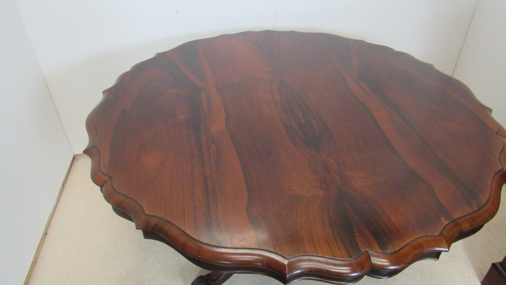 A 19th-century circular rosewood tilt-top table displayed in an upright position, featuring a shaped scalloped rim, prominent book-matched timber grain, and a carved pedestal base. View V