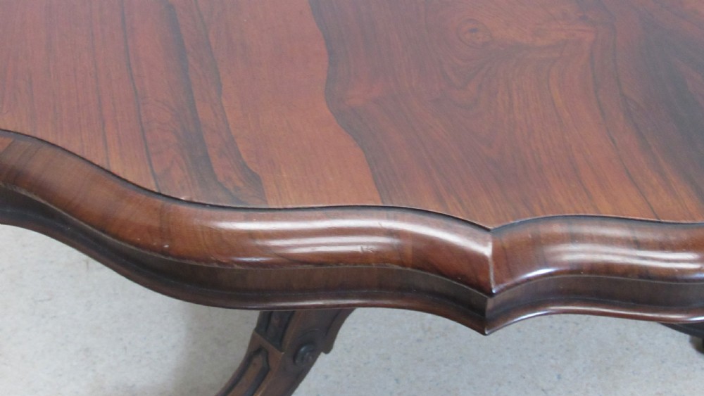 A 19th-century circular rosewood tilt-top table displayed in an upright position, featuring a shaped scalloped rim, prominent book-matched timber grain, and a carved pedestal base. View VI
