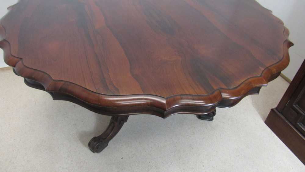 A 19th-century circular rosewood tilt-top table displayed in an upright position, featuring a shaped scalloped rim, prominent book-matched timber grain, and a carved pedestal base. View XIV