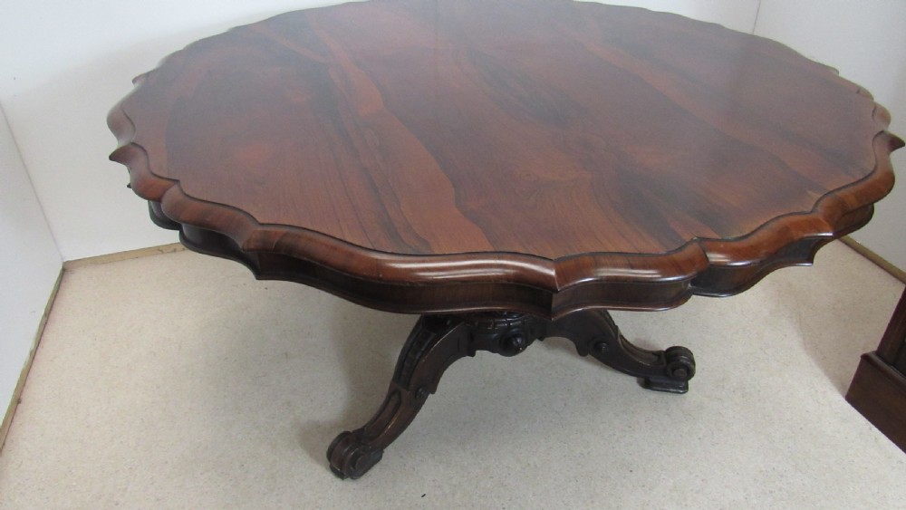 A 19th-century circular rosewood tilt-top table displayed in an upright position, featuring a shaped scalloped rim, prominent book-matched timber grain, and a carved pedestal base. View XV