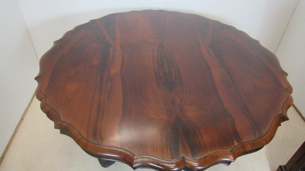 A 19th-century circular rosewood tilt-top table displayed in an upright position, featuring a shaped scalloped rim, prominent book-matched timber grain, and a carved pedestal base. View XVI