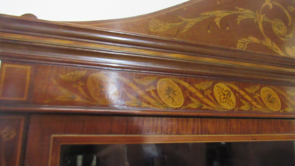 An antique mahogany glazed display cabinet featuring marquetry inlay, two glass doors, four small drawers, and square tapered legs. View V
