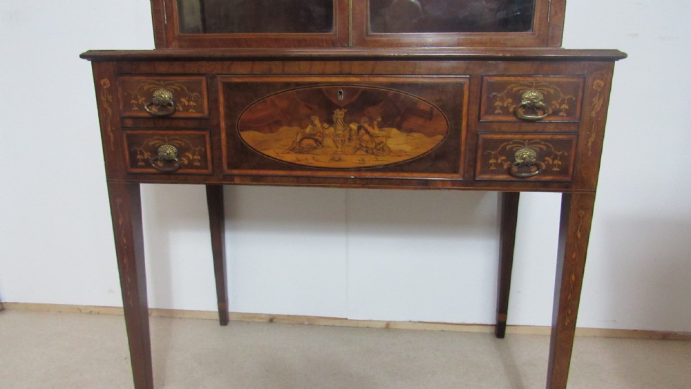 An antique mahogany glazed display cabinet featuring marquetry inlay, two glass doors, four small drawers, and square tapered legs. View VI