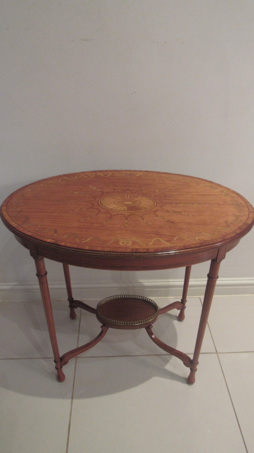 An oval antique satinwood occasional table featuring decorative marquetry inlay on top, tapered legs, and a small lower shelf with a brass gallery rail. View II