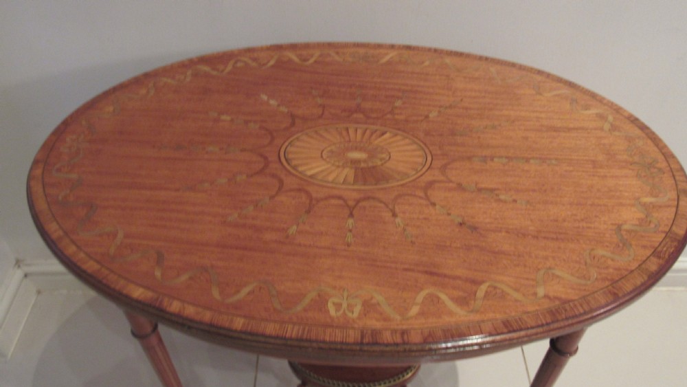 An oval antique satinwood occasional table featuring decorative marquetry inlay on top, tapered legs, and a small lower shelf with a brass gallery rail. View III