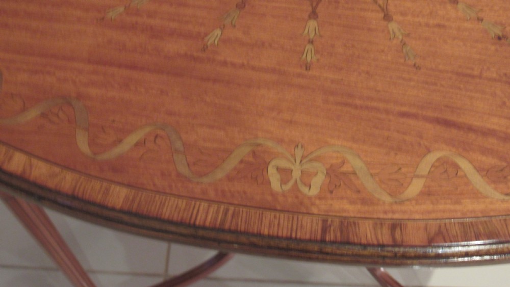 An oval antique satinwood occasional table featuring decorative marquetry inlay on top, tapered legs, and a small lower shelf with a brass gallery rail. View VI