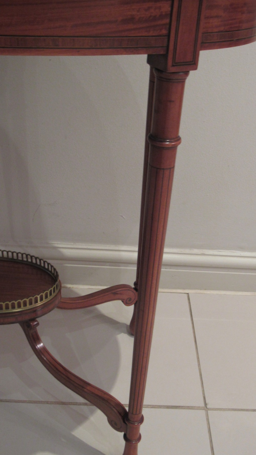 An oval antique satinwood occasional table featuring decorative marquetry inlay on top, tapered legs, and a small lower shelf with a brass gallery rail. View X
