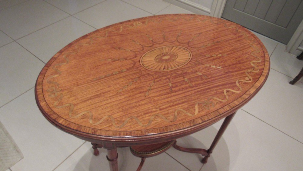 An oval antique satinwood occasional table featuring decorative marquetry inlay on top, tapered legs, and a small lower shelf with a brass gallery rail. View XIII