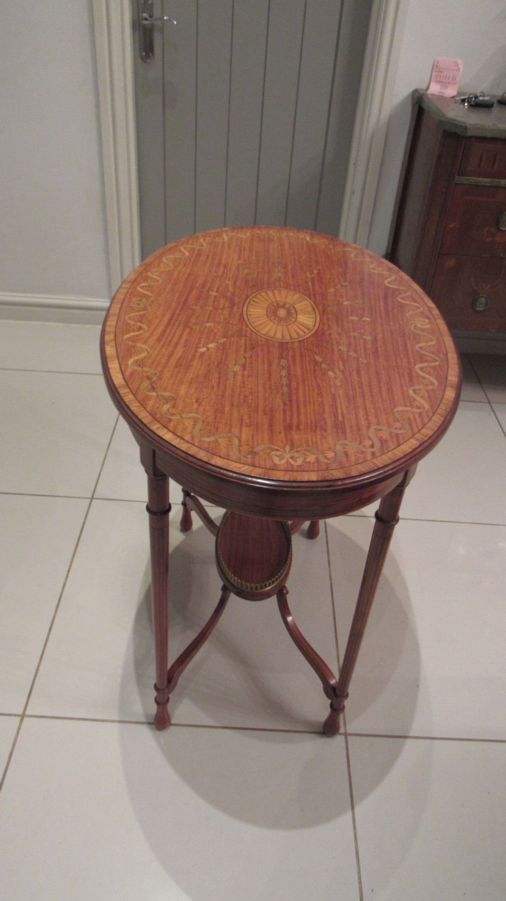 An oval antique satinwood occasional table featuring decorative marquetry inlay on top, tapered legs, and a small lower shelf with a brass gallery rail. View XV