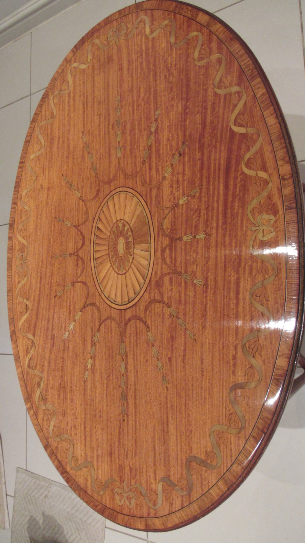 An oval antique satinwood occasional table featuring decorative marquetry inlay on top, tapered legs, and a small lower shelf with a brass gallery rail. View XIX