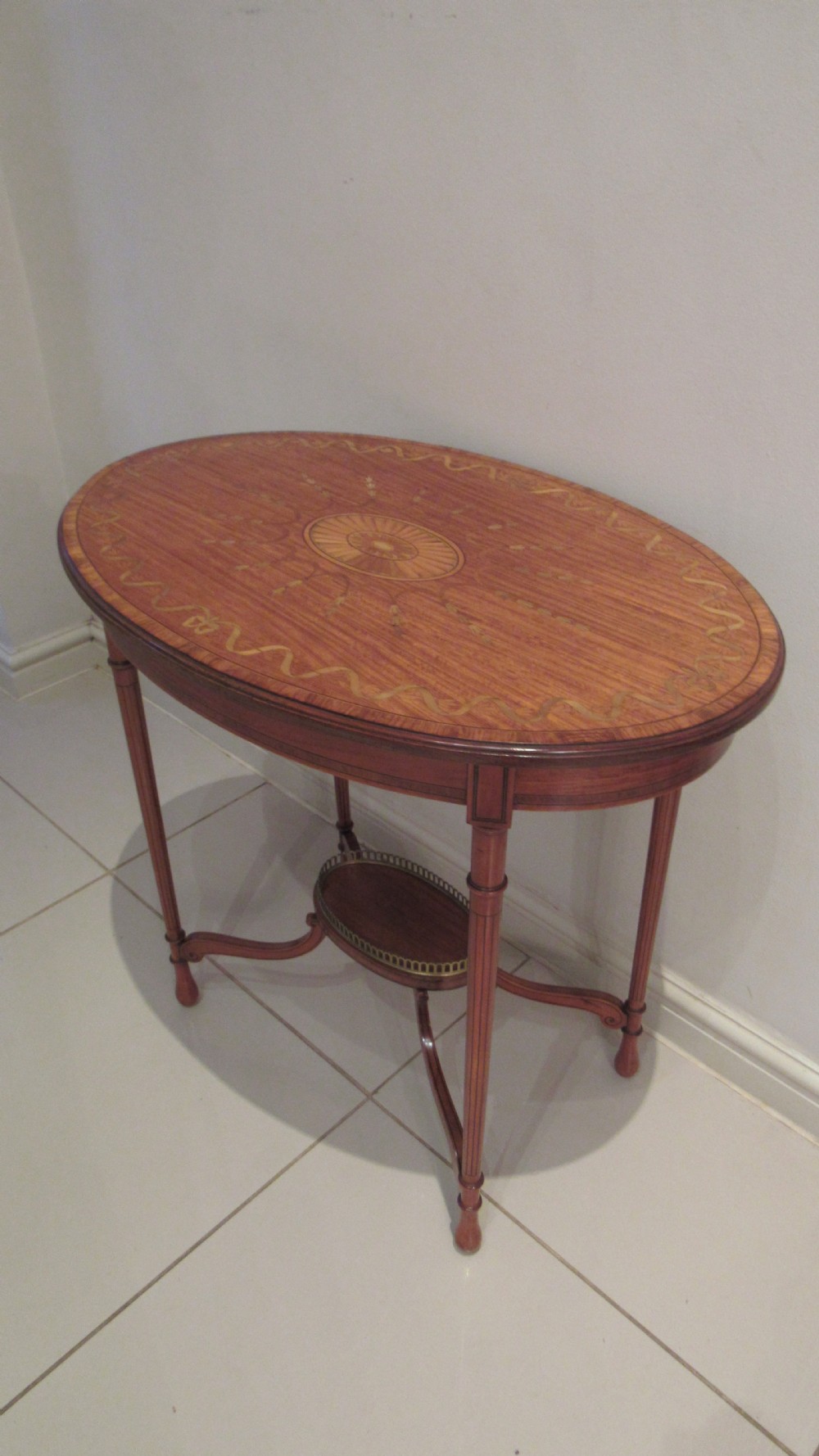 An oval antique satinwood occasional table featuring decorative marquetry inlay on top, tapered legs, and a small lower shelf with a brass gallery rail. View XX