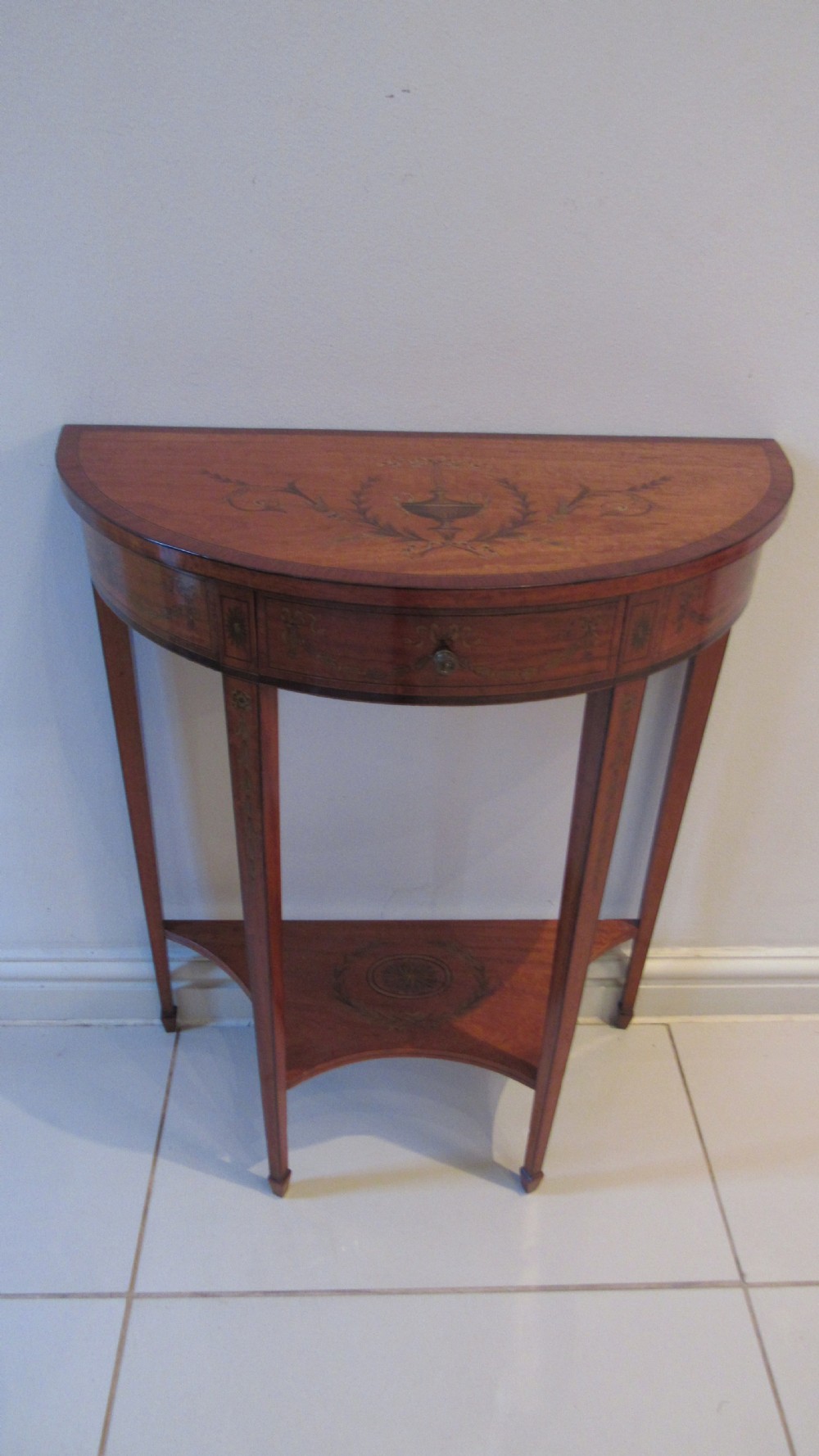 A semi-circular golden satinwood demilune console table featuring urn marquetry inlay, slender tapered legs, and an under-tier shelf. View II