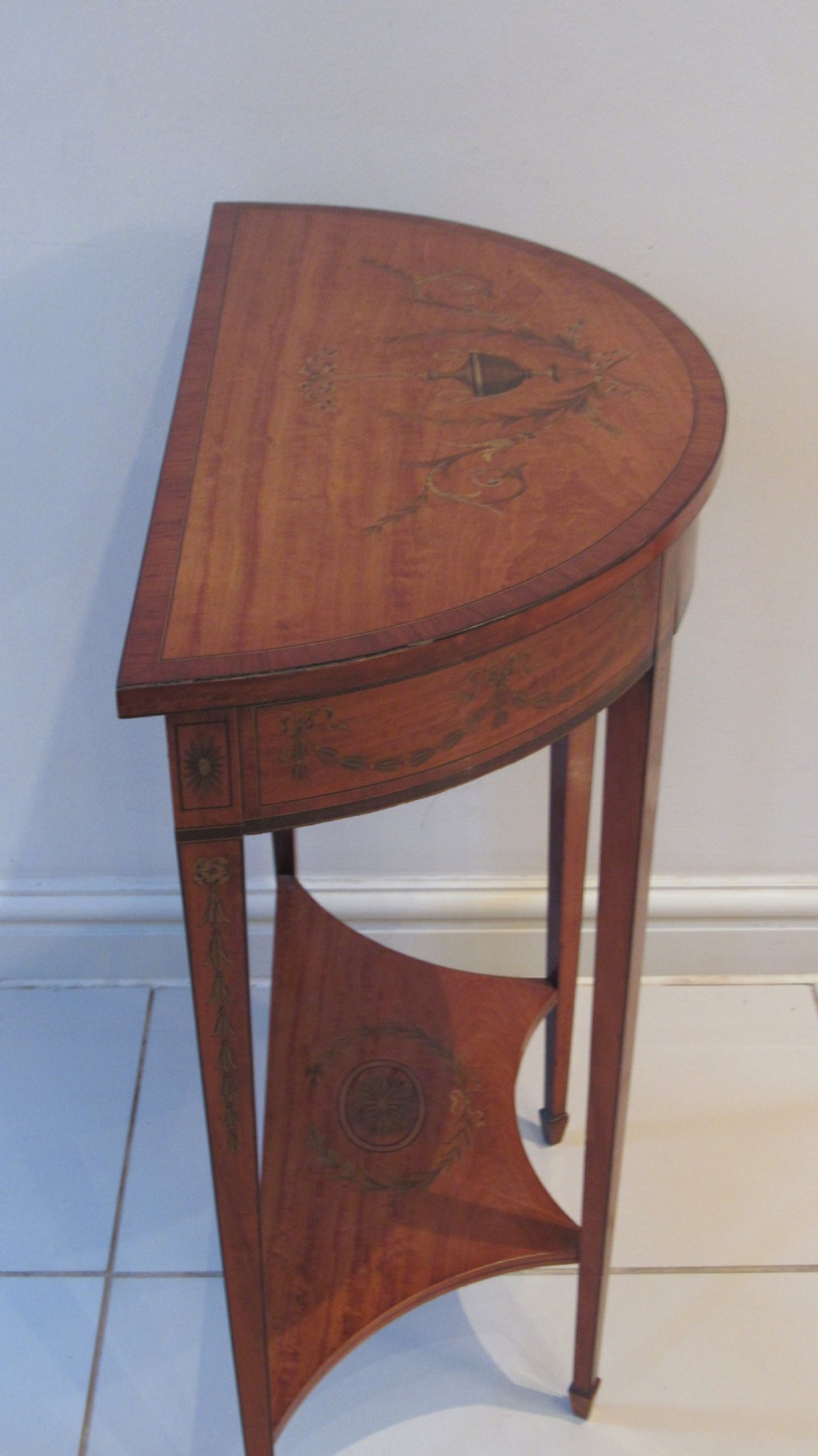 A semi-circular golden satinwood demilune console table featuring urn marquetry inlay, slender tapered legs, and an under-tier shelf. View XII