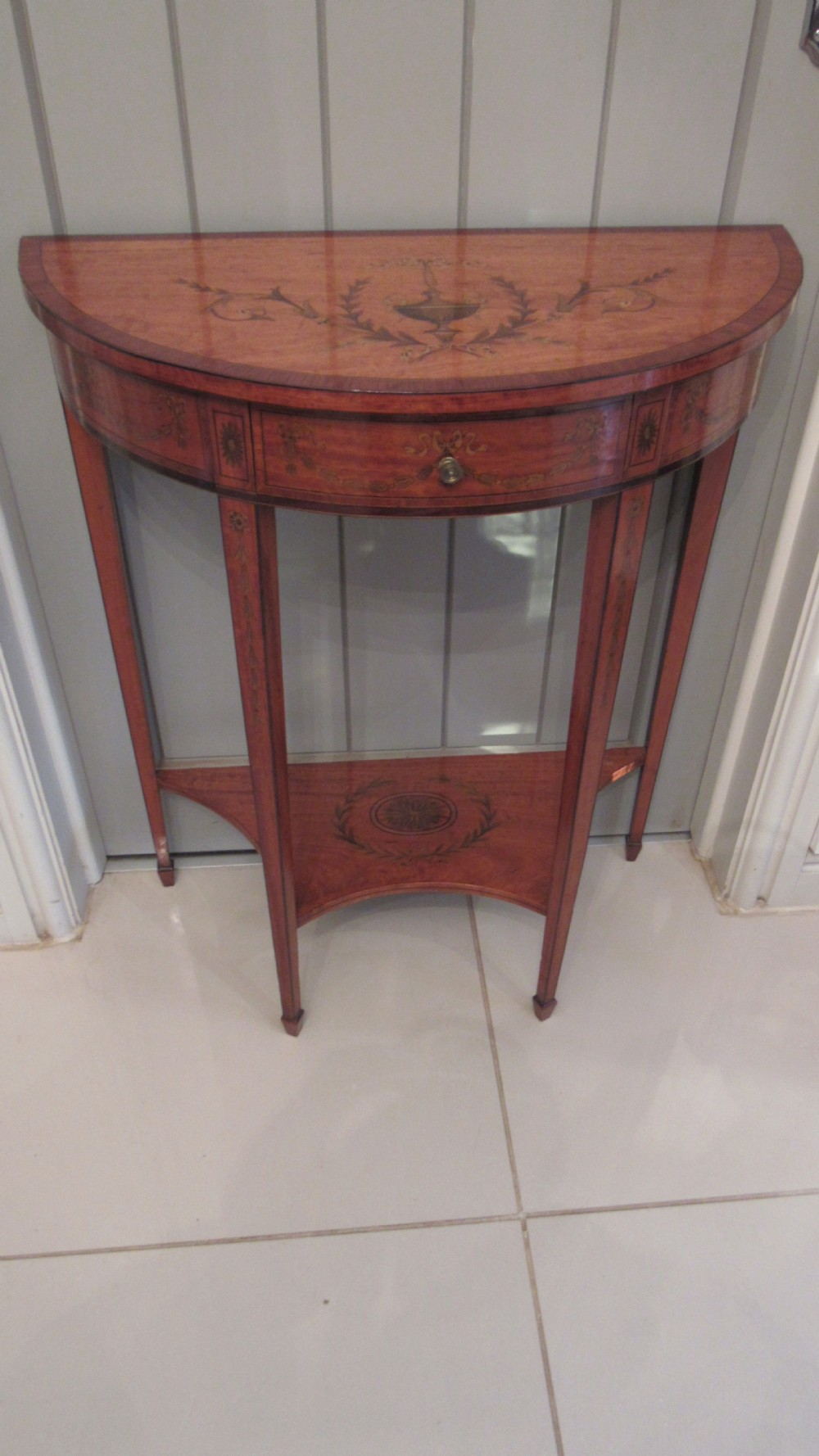A semi-circular golden satinwood demilune console table featuring urn marquetry inlay, slender tapered legs, and an under-tier shelf. View XVI