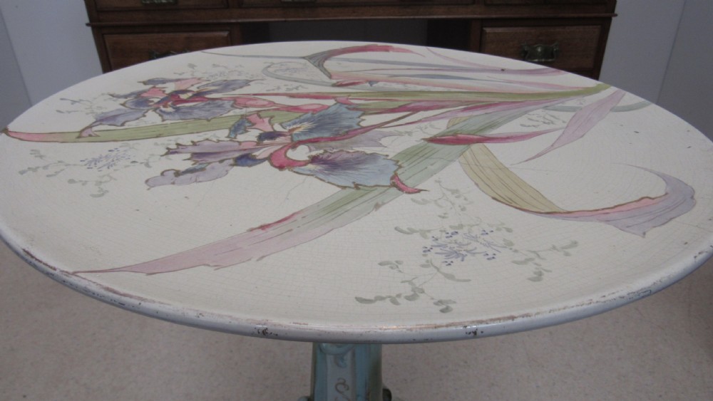 A circular Art Nouveau pottery table featuring a hand-painted floral iris design on a cream-glazed top supported by a sculpted pedestal base. View XVI