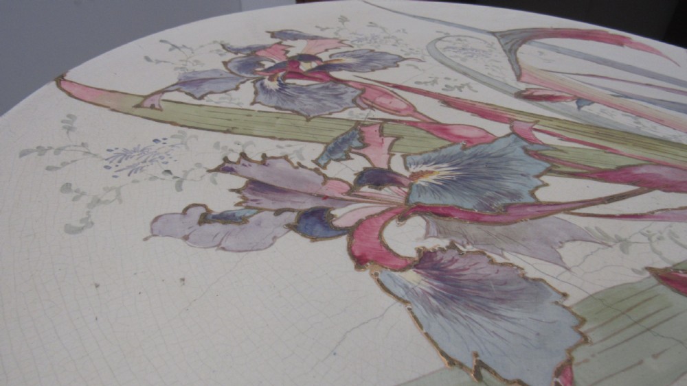 A circular Art Nouveau pottery table featuring a hand-painted floral iris design on a cream-glazed top supported by a sculpted pedestal base. View XVII