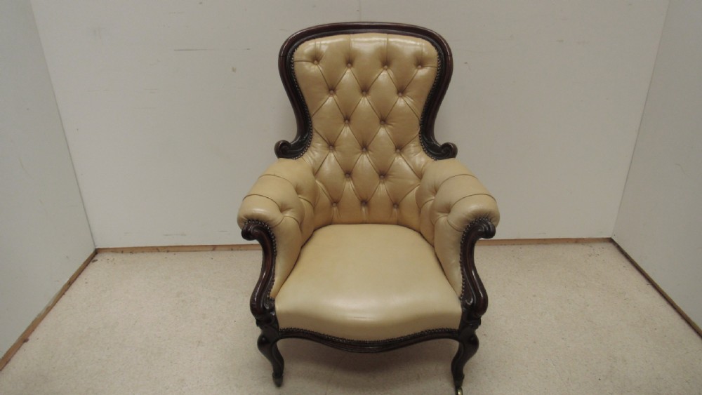 A Victorian library armchair with deep-buttoned cream leather upholstery, a carved dark mahogany frame, padded armrests, and cabriole front legs with brass castors.