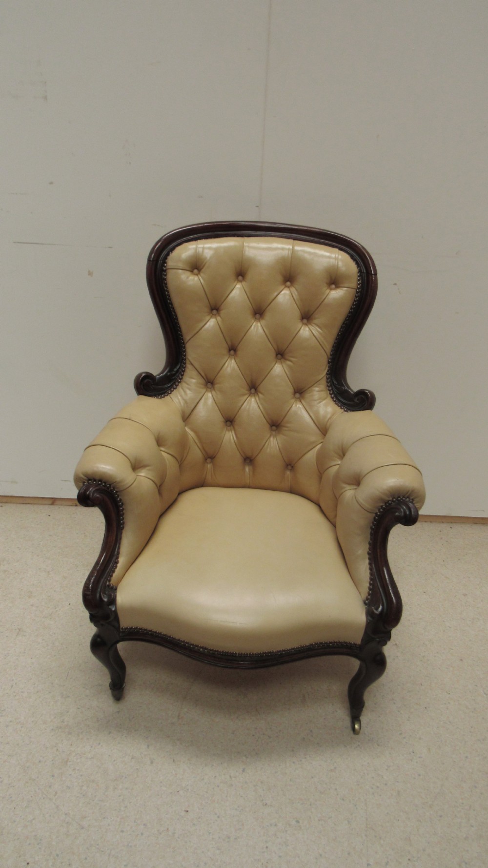 A Victorian library armchair with deep-buttoned cream leather upholstery, a carved dark mahogany frame, padded armrests, and cabriole front legs with brass castors. View II