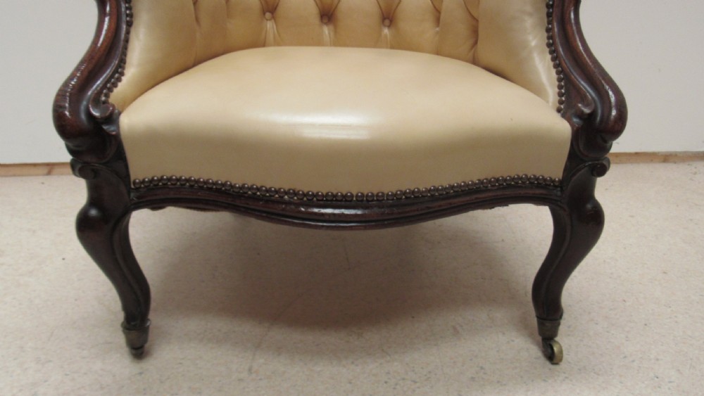 A Victorian library armchair with deep-buttoned cream leather upholstery, a carved dark mahogany frame, padded armrests, and cabriole front legs with brass castors. View VII
