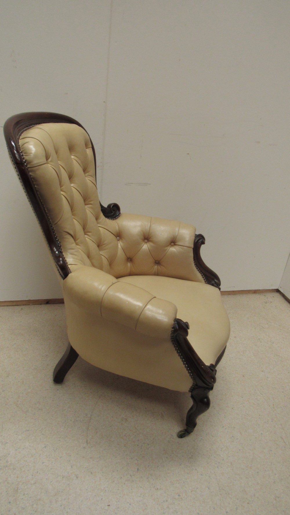 A Victorian library armchair with deep-buttoned cream leather upholstery, a carved dark mahogany frame, padded armrests, and cabriole front legs with brass castors. View XII