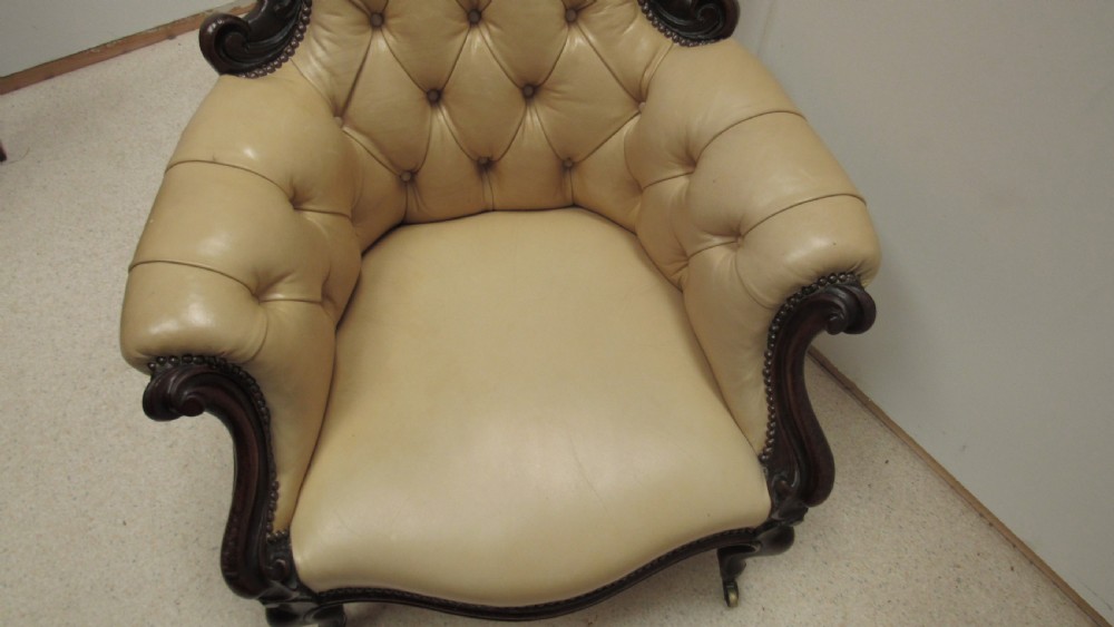 A Victorian library armchair with deep-buttoned cream leather upholstery, a carved dark mahogany frame, padded armrests, and cabriole front legs with brass castors. View XV