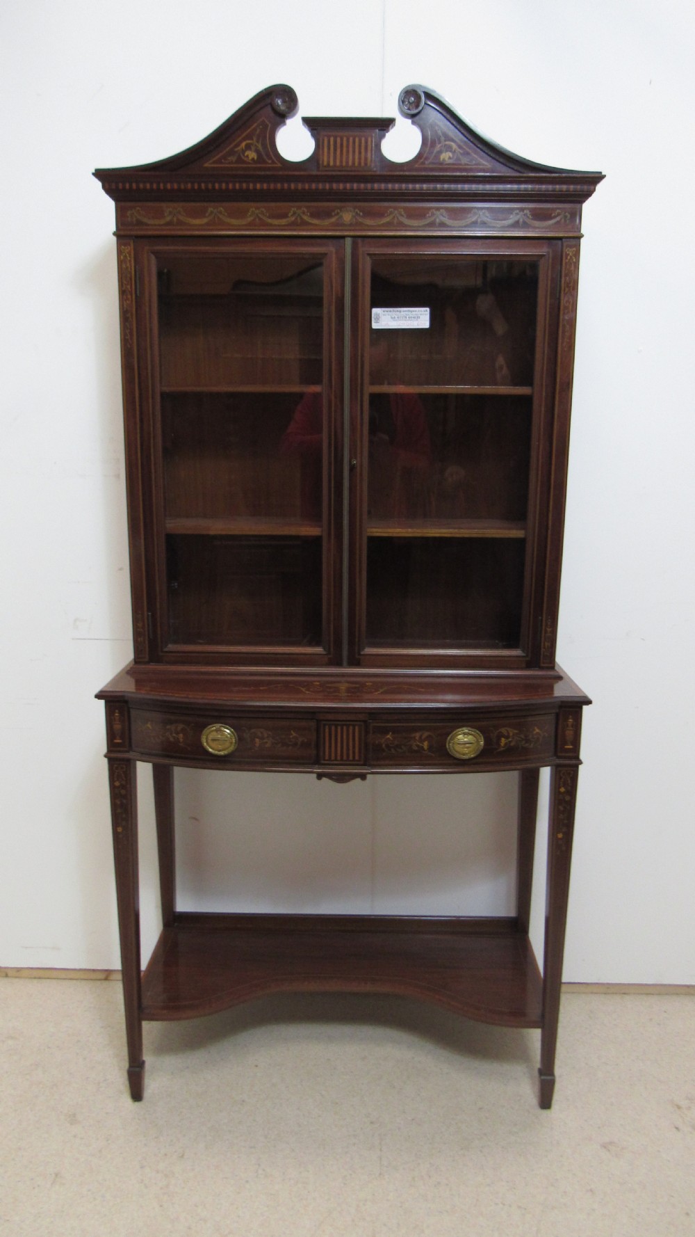 An Edwardian dark mahogany display cabinet featuring a swan-neck pediment, delicate satinwood floral marquetry, two glazed upper doors, drawers with brass handles, and tapered legs with an undertier shelf.