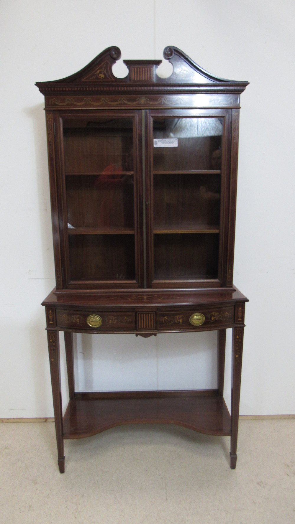 An Edwardian dark mahogany display cabinet featuring a swan-neck pediment, delicate satinwood floral marquetry, two glazed upper doors, drawers with brass handles, and tapered legs with an undertier shelf. View III