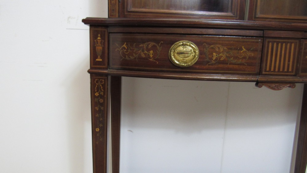 An Edwardian dark mahogany display cabinet featuring a swan-neck pediment, delicate satinwood floral marquetry, two glazed upper doors, drawers with brass handles, and tapered legs with an undertier shelf. View IV