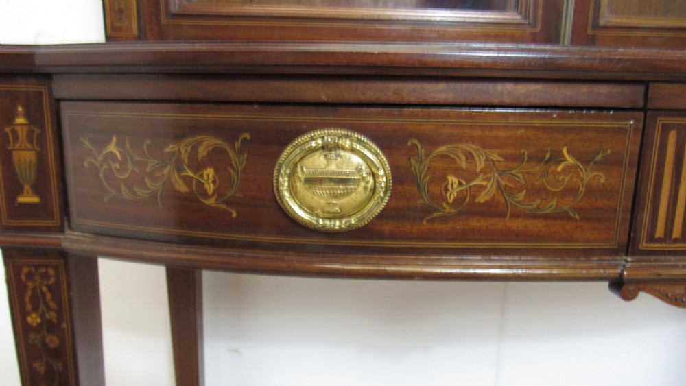 An Edwardian dark mahogany display cabinet featuring a swan-neck pediment, delicate satinwood floral marquetry, two glazed upper doors, drawers with brass handles, and tapered legs with an undertier shelf. View IX