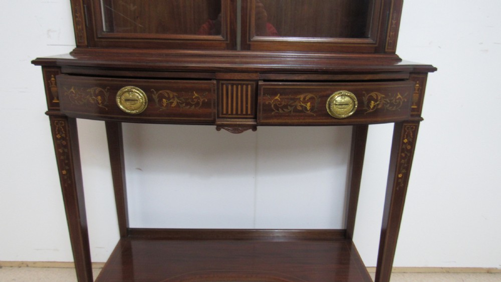 An Edwardian dark mahogany display cabinet featuring a swan-neck pediment, delicate satinwood floral marquetry, two glazed upper doors, drawers with brass handles, and tapered legs with an undertier shelf. View X