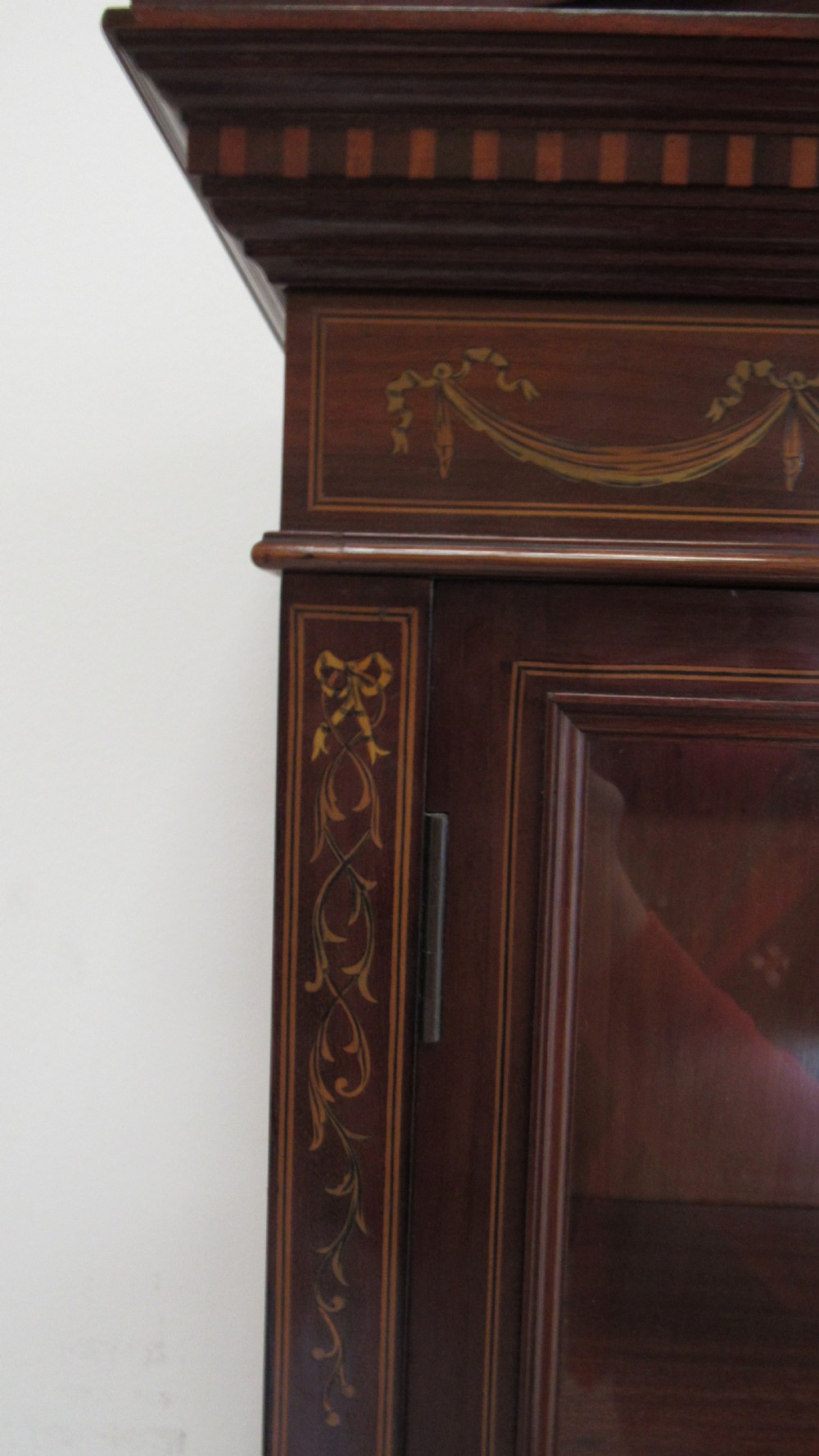 An Edwardian dark mahogany display cabinet featuring a swan-neck pediment, delicate satinwood floral marquetry, two glazed upper doors, drawers with brass handles, and tapered legs with an undertier shelf. View XV