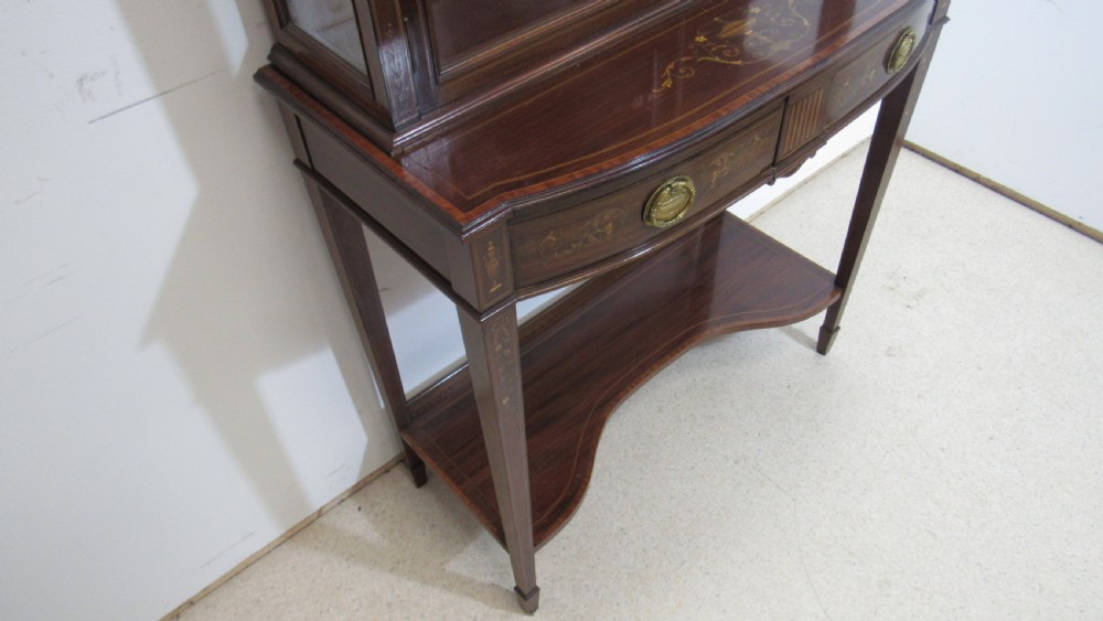 An Edwardian dark mahogany display cabinet featuring a swan-neck pediment, delicate satinwood floral marquetry, two glazed upper doors, drawers with brass handles, and tapered legs with an undertier shelf. View XVI