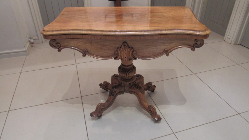 An antique Victorian walnut card table featuring a serpentine fold-over top resting on a carved central pedestal and scroll feet.