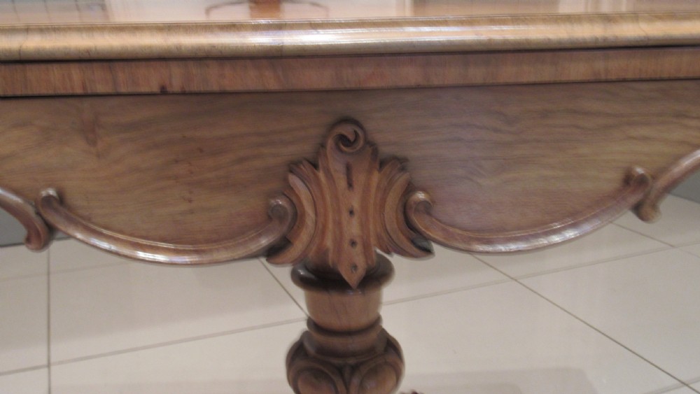 An antique Victorian walnut card table featuring a serpentine fold-over top resting on a carved central pedestal and scroll feet. View VIII