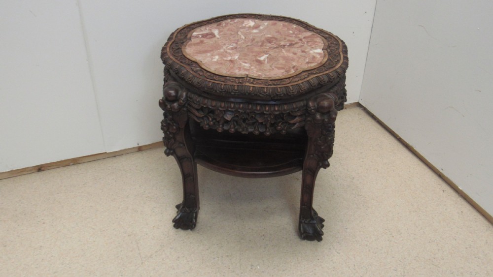 A circular dark wood urn stand featuring an inset pink marble top, detailed relief carving on the apron, curved cabriole legs, and a lower shelf.
