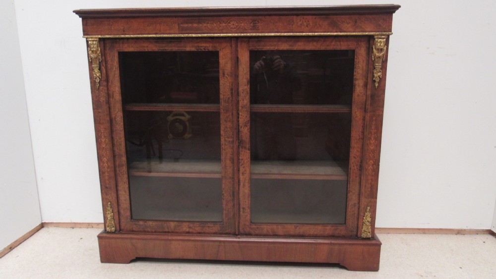 A 19th-century Victorian burr walnut pier cabinet featuring floral marquetry inlay, glazed double doors, glass shelves, and gilt brass corner mounts.