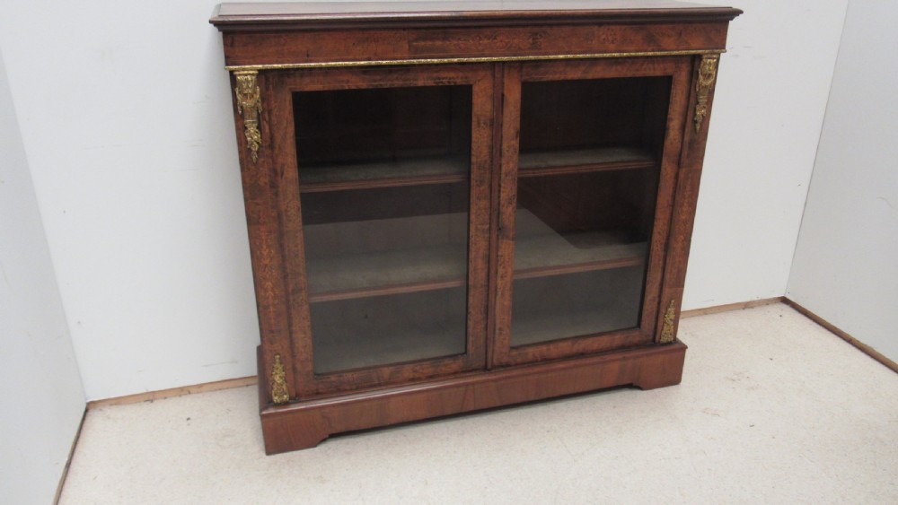 A 19th-century Victorian burr walnut pier cabinet featuring floral marquetry inlay, glazed double doors, glass shelves, and gilt brass corner mounts. View X