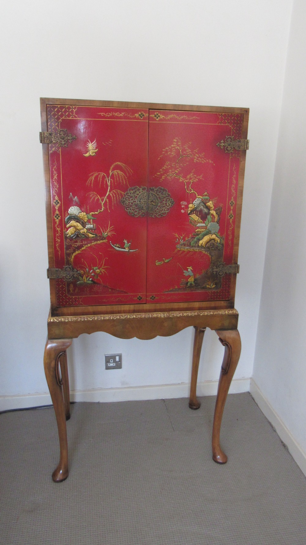 A red lacquered chinoiserie cocktail cabinet decorated with gilt landscapes and figures, featuring ornate brass hinges and set on a carved timber stand with cabriole legs.