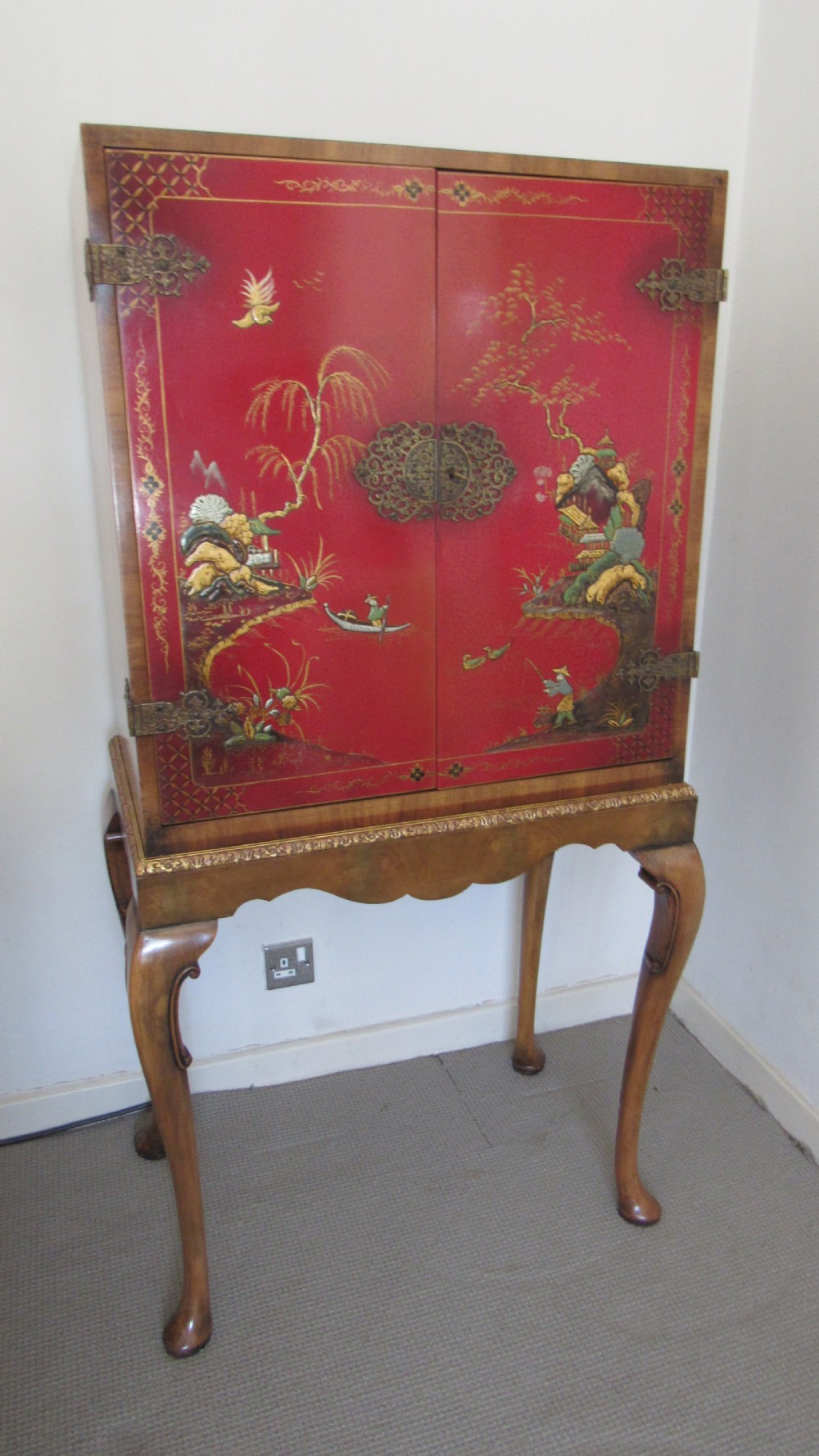 A red lacquered chinoiserie cocktail cabinet decorated with gilt landscapes and figures, featuring ornate brass hinges and set on a carved timber stand with cabriole legs. View II