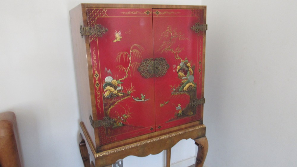 A red lacquered chinoiserie cocktail cabinet decorated with gilt landscapes and figures, featuring ornate brass hinges and set on a carved timber stand with cabriole legs. View V