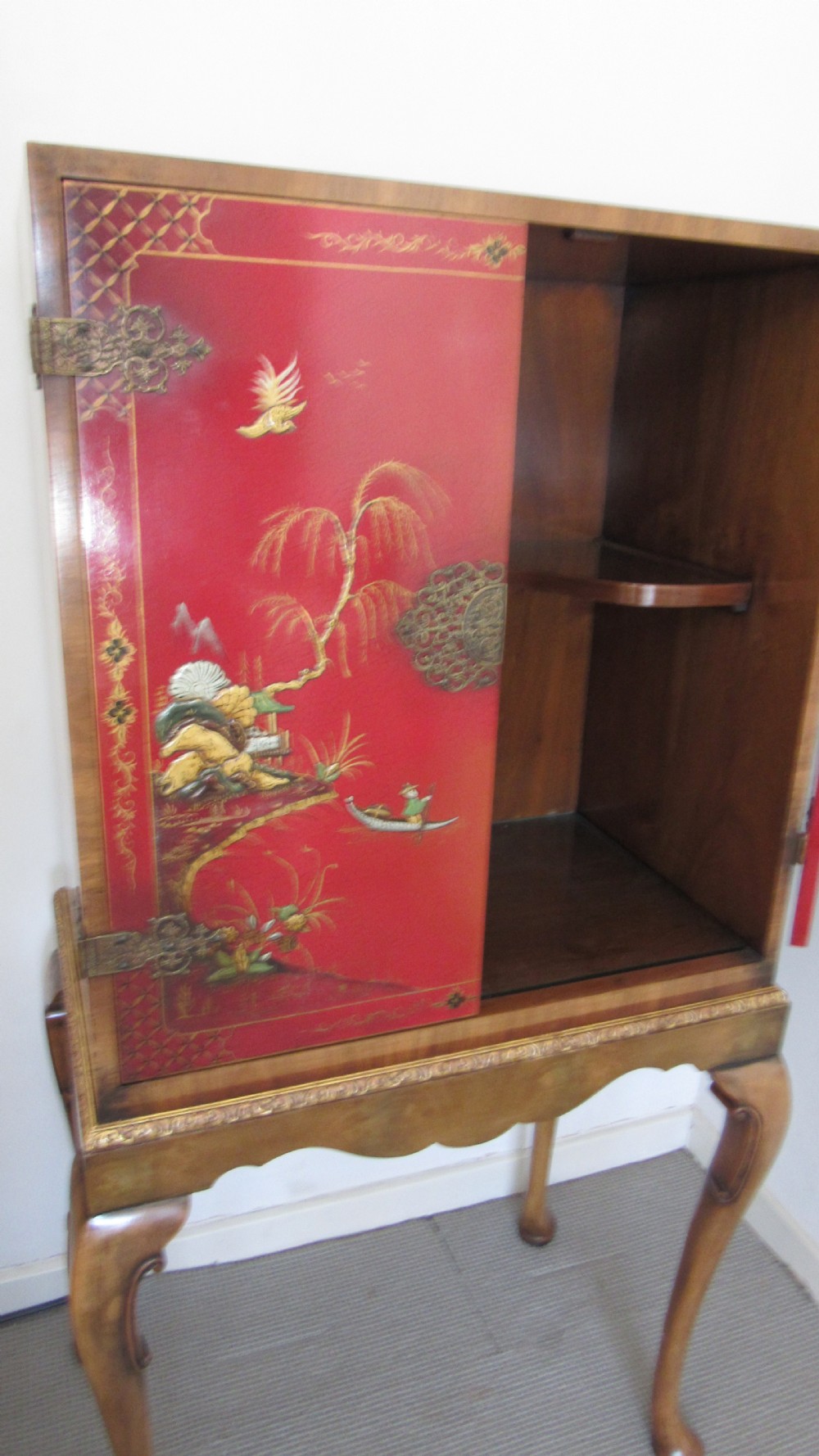 A red lacquered chinoiserie cocktail cabinet decorated with gilt landscapes and figures, featuring ornate brass hinges and set on a carved timber stand with cabriole legs. View XV