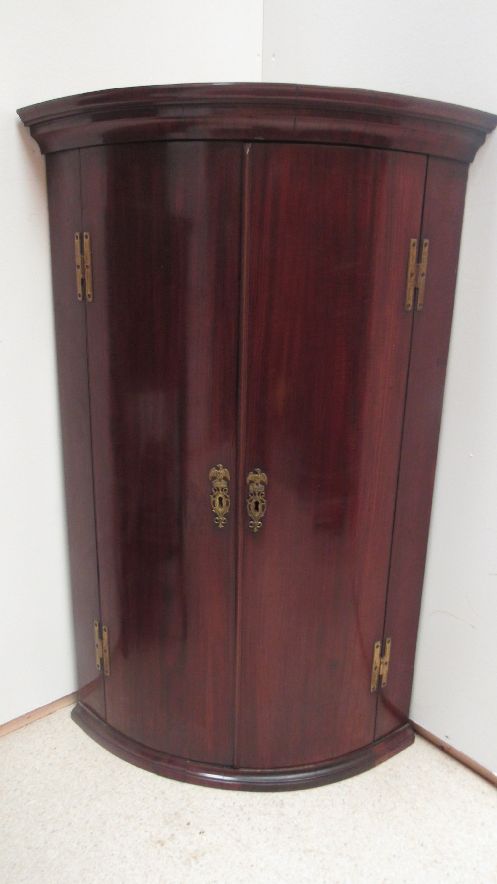 An antique Georgian bowfront corner cupboard crafted from dark mahogany with twin doors, brass H-hinges, and decorative escutcheons.